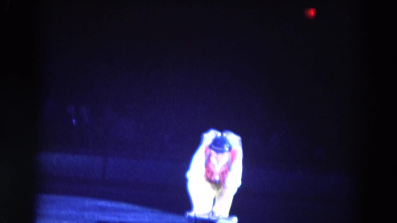 Male performer in red vest and bowler hat crouches on an ice rink under blue stage light.