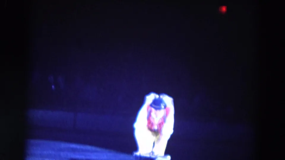 In 1969 Chicago home-movie footage, a male ice performer in a red vest and bowler hat bends low under blue rink lighting.