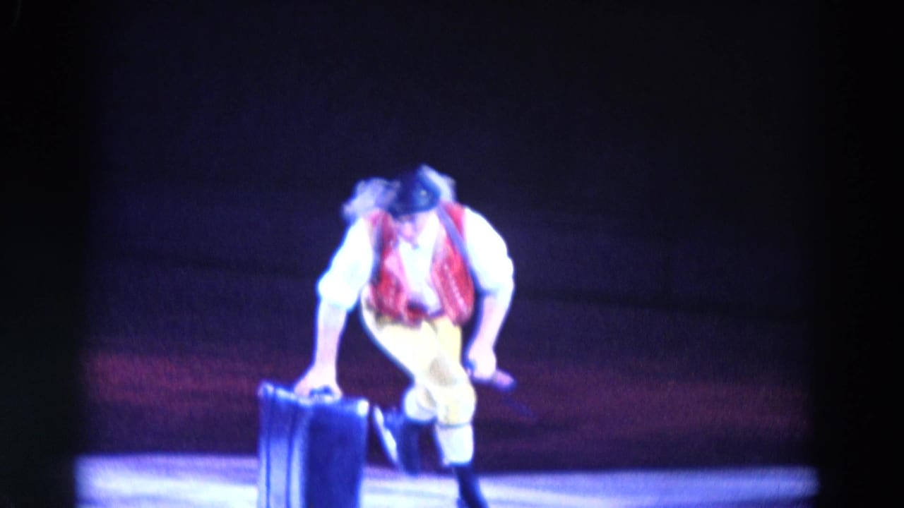 A male ice performer in a red vest and bowler hat leans toward a dark suitcase prop during the 1969 Chicago routine.