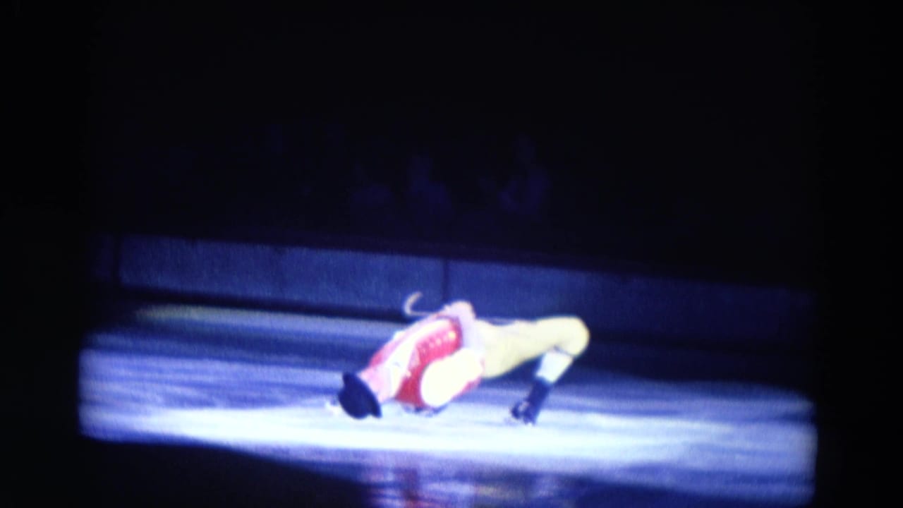 Male ice performer in red vest and yellow tights bends backward on a lit rink.