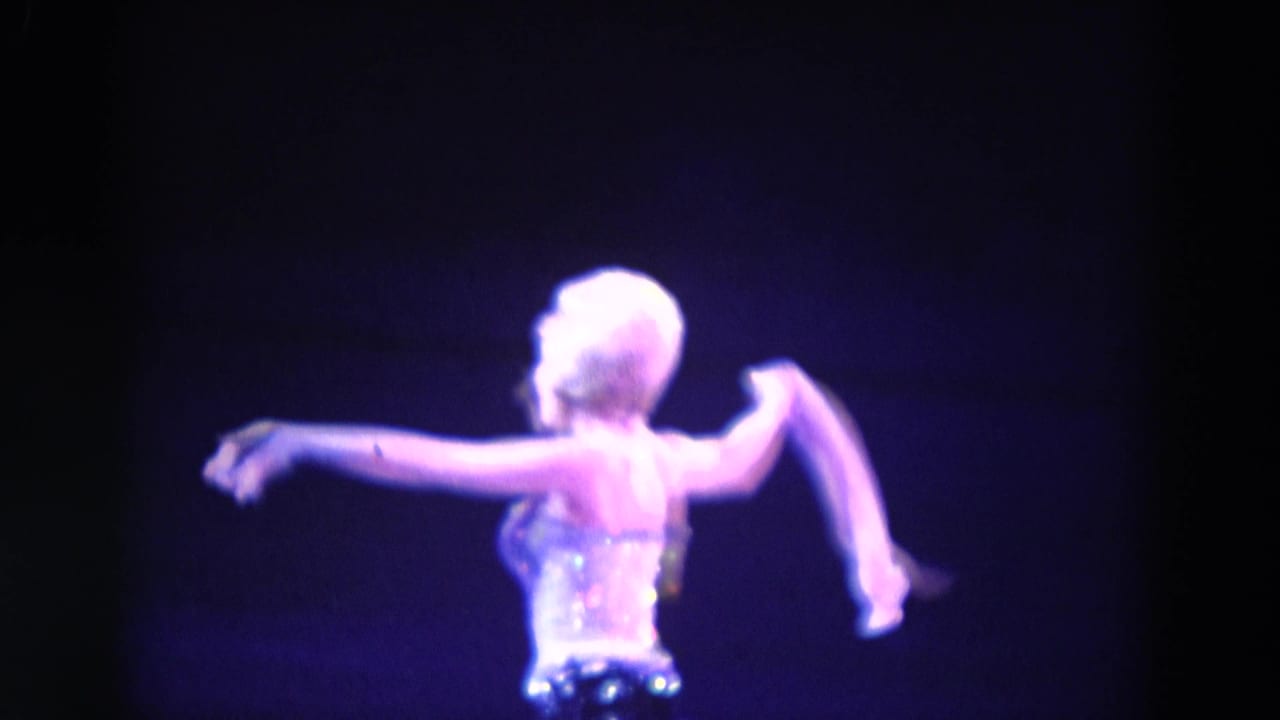 In 1969 Chicago home-movie footage, a woman in a sparkling costume performs a solo dance under purple stage light.