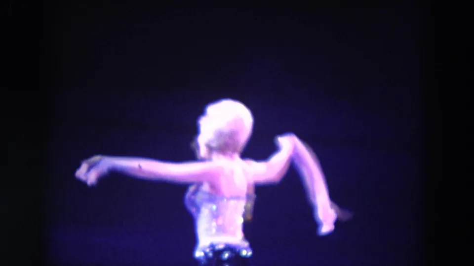 In 1969 Chicago home-movie footage, a woman in a sparkling costume performs a solo dance under purple stage light.