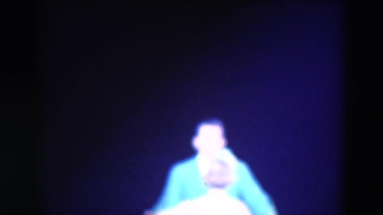 A man in a bright blue shirt appears on the dark stage, with another performer partly visible in the foreground.
