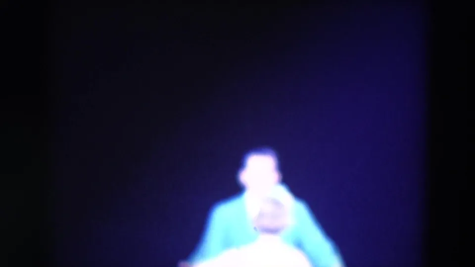 A man in a bright blue shirt appears on the dark stage, with another performer partly visible in the foreground.