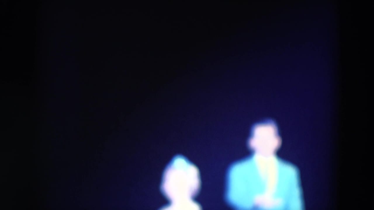 1969 Chicago 8mm frame showing a softly focused man in a bright blue jacket and a woman in a pale dress on a dim stage.