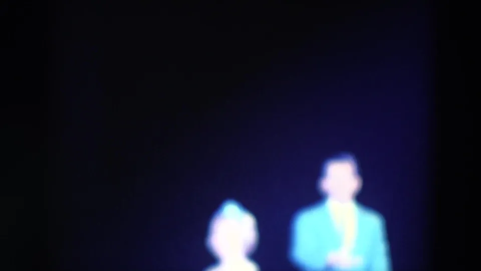 1969 Chicago 8mm frame showing a softly focused man in a bright blue jacket and a woman in a pale dress on a dim stage.