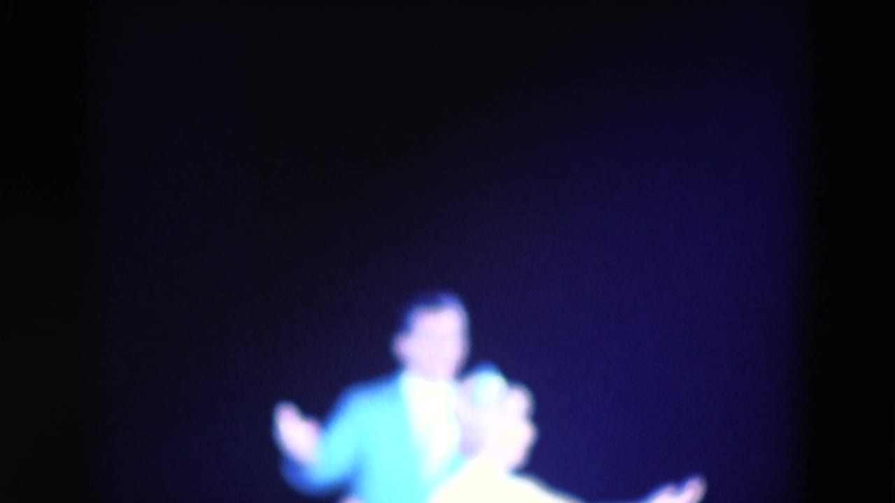 1969 Chicago home-movie frame of the male performer gesturing while the woman stands nearby in blue-purple stage light.