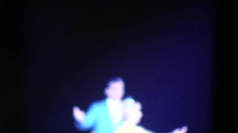 1969 Chicago home-movie frame of the male performer gesturing while the woman stands nearby in blue-purple stage light.