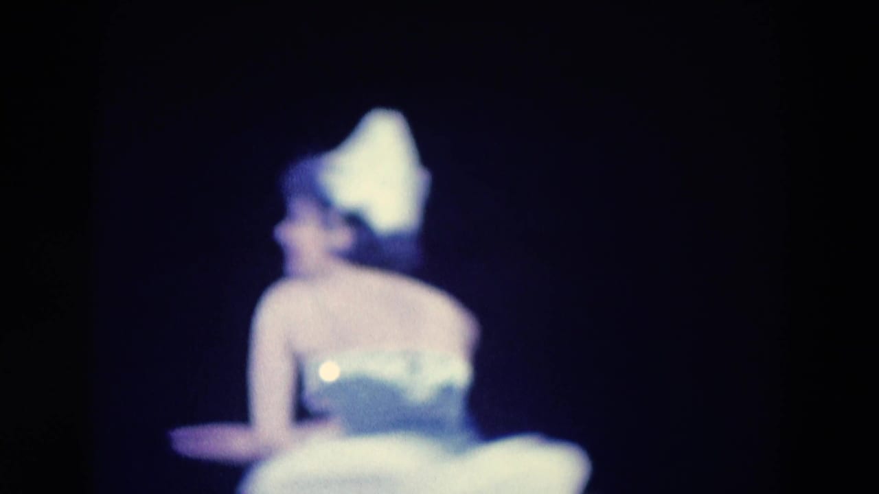 Ballerina in a white tutu and pointed hat turned partly away against a dark backdrop.