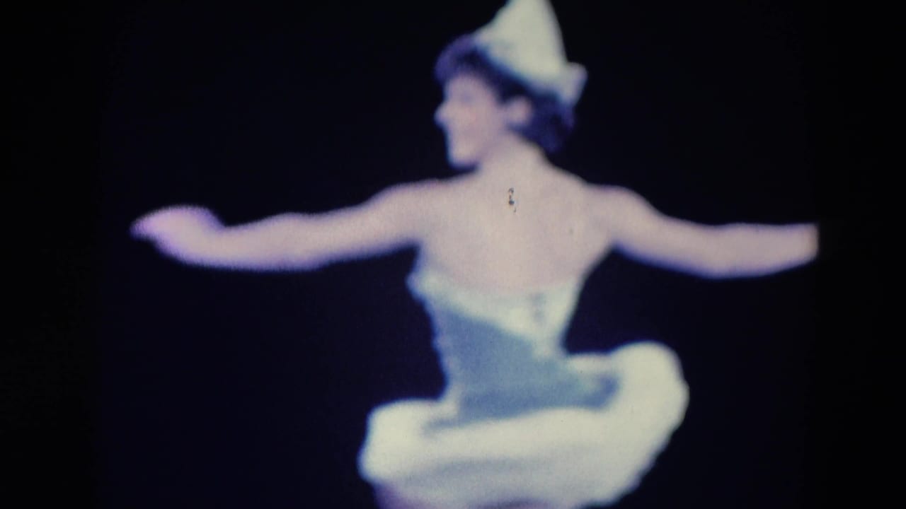 Ballerina seen from behind with both arms extended against a dark background.
