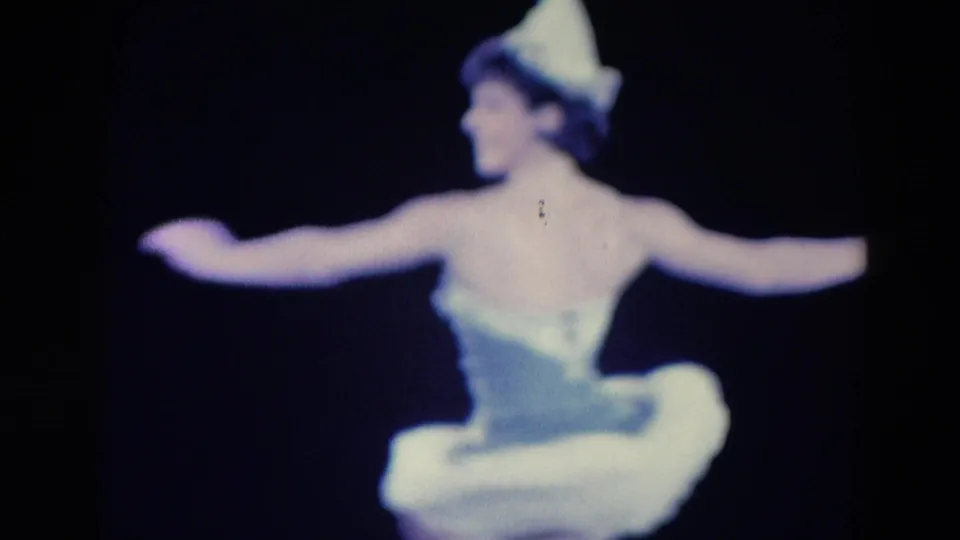 1969 Chicago 8mm frame showing the ballerina from behind with both arms extended, her white tutu and pointed hat visible against a dark stage-like background.