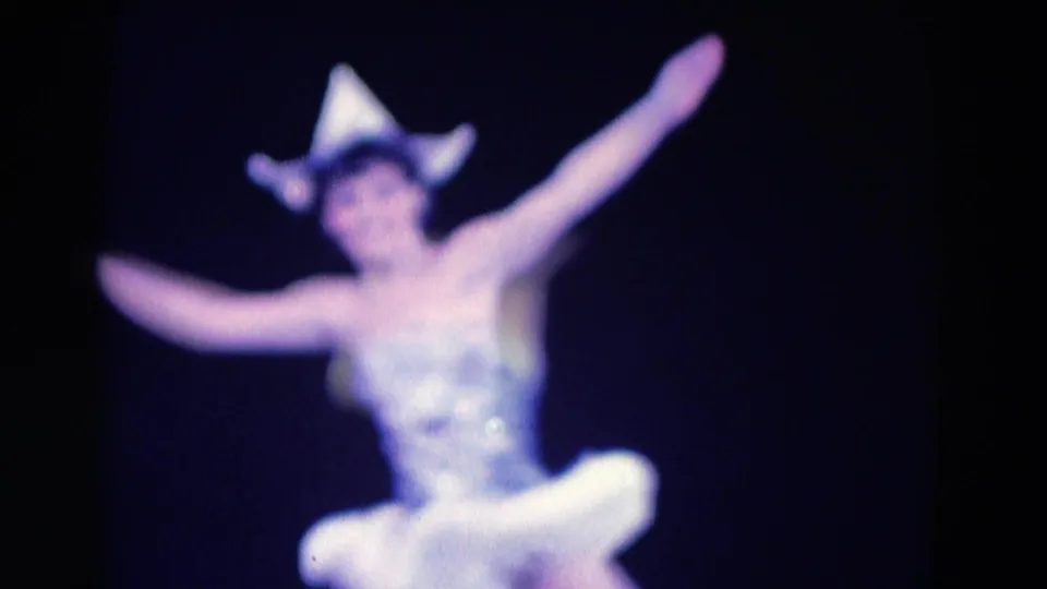 Soft 1969 Chicago 8mm frame showing the ballerina facing the camera with arms spread upward, wearing a white tutu and pointed hat during the performance.