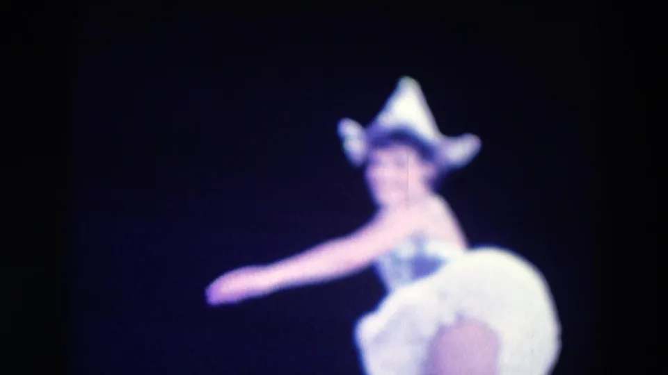 A ballerina in a white tutu and bright headpiece turns with one arm extended in 1969 Chicago 8mm footage.