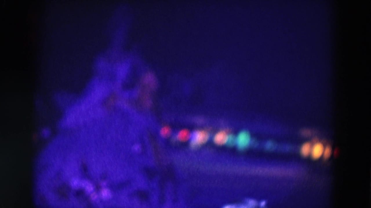 1969 Chicago 8mm frame with a blurred figure skater under saturated purple light and small colored lights across the background.