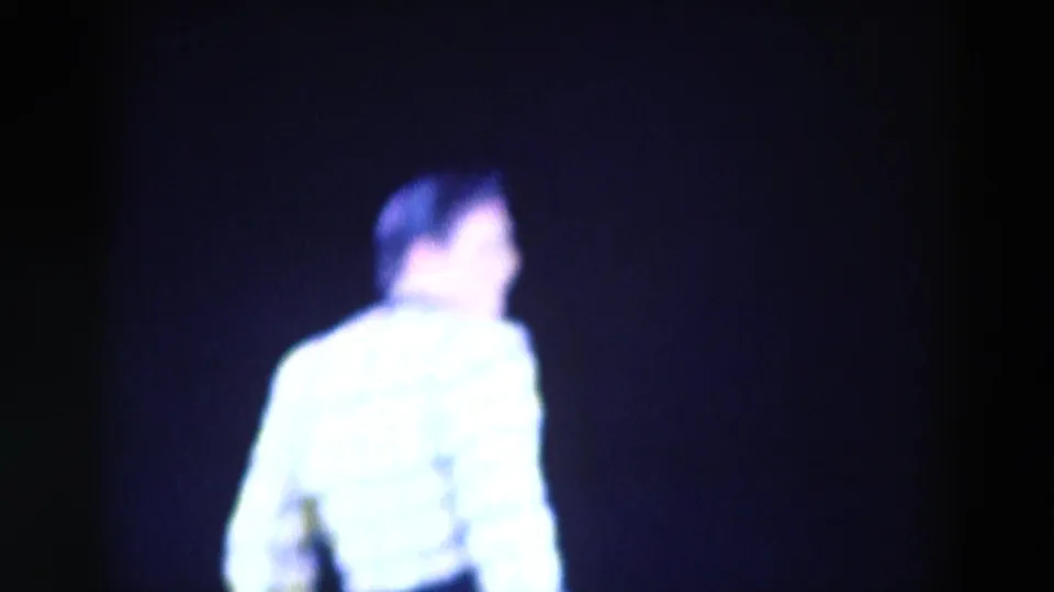 1969 Chicago 8mm frame showing a dark-haired skater in a bright white outfit, viewed from behind in blue stage light.