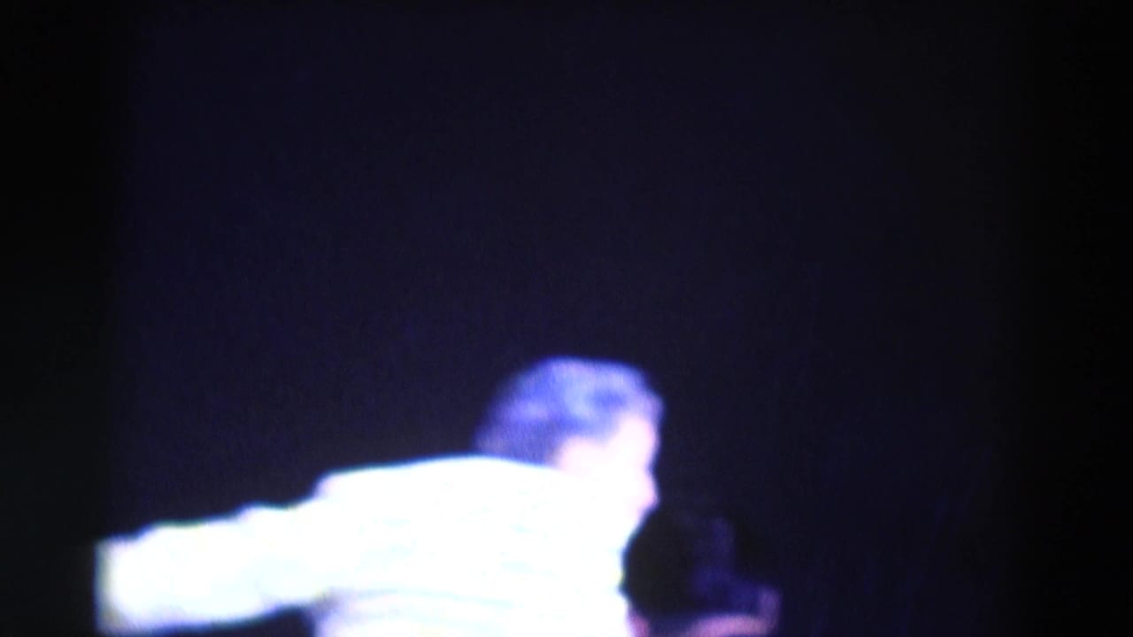 Blurred performer in a bright shirt moving across a dark stage.
