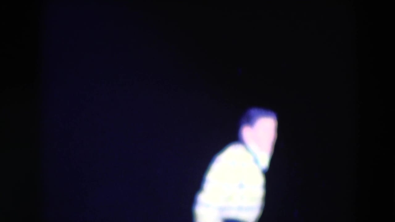 Brightly lit performer in a patterned jacket leans on a dark stage.
