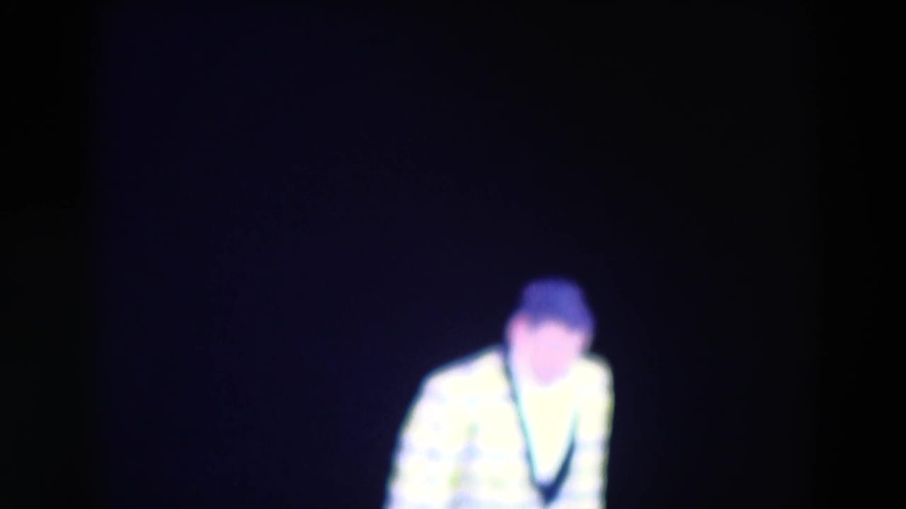 Performer in a bright jacket crouches or bends under stage lighting.
