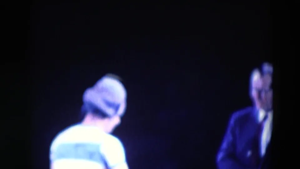 A performer seen from behind appears to face a man in a dark suit, suggesting a staged exchange in the Chicago comedy footage.
