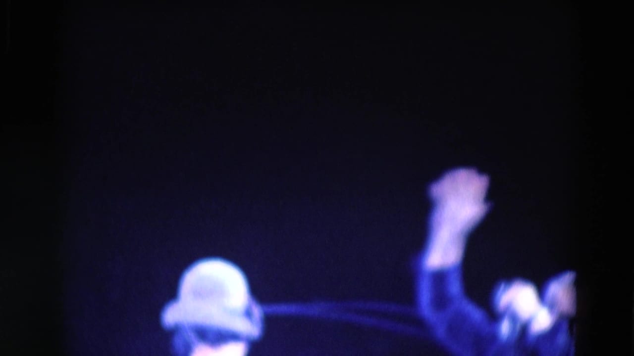 Blurred stage view with a helmeted or capped figure at left and a raised arm near center.