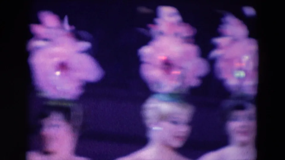 Soft 1969 Chicago 8mm frame showing performers in large pink feathered hats and sequined stage costumes.
