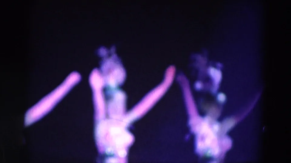 Low-light 8mm frame of women dancers in feathered headpieces and bright costumes moving through a stage routine.