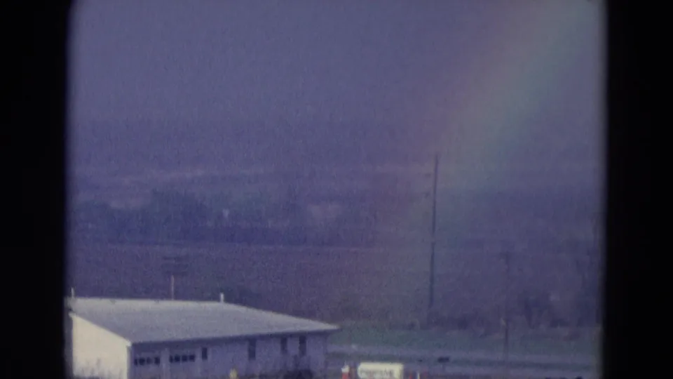 1969 8mm frame showing a faint rainbow above open land, with a small light-colored building and distant landscape visible.