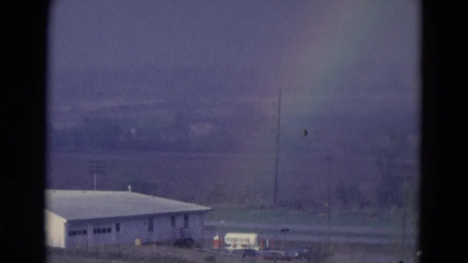 1969 8mm frame from the Chicago, Illinois area showing a faint rainbow over a white roadside building, poles, and distant trees.