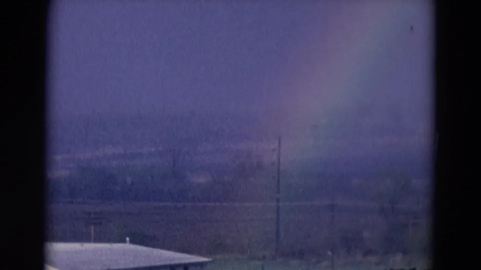 1969 Chicago-area 8mm frame showing the rainbow over a white building roofline, bare trees, utility pole, and distant open land.