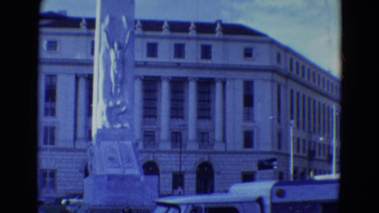 Tall sculpted monument in front of a columned classical building with a partial vehicle at the street edge.