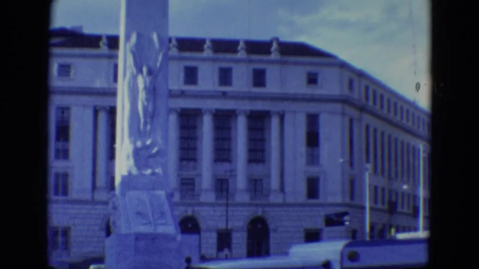 Street-level 1969 home-movie view in Diller, Nebraska, with a sculpted monument foregrounding a large classical building facade.