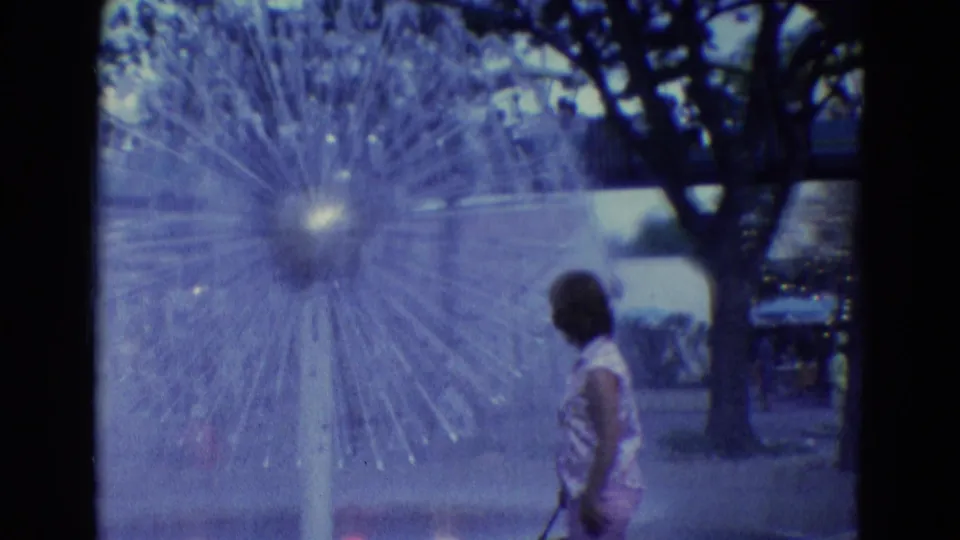 1969 Diller, Nebraska frame of a woman in a patterned dress near a starburst-style outdoor fountain and shaded walkway.