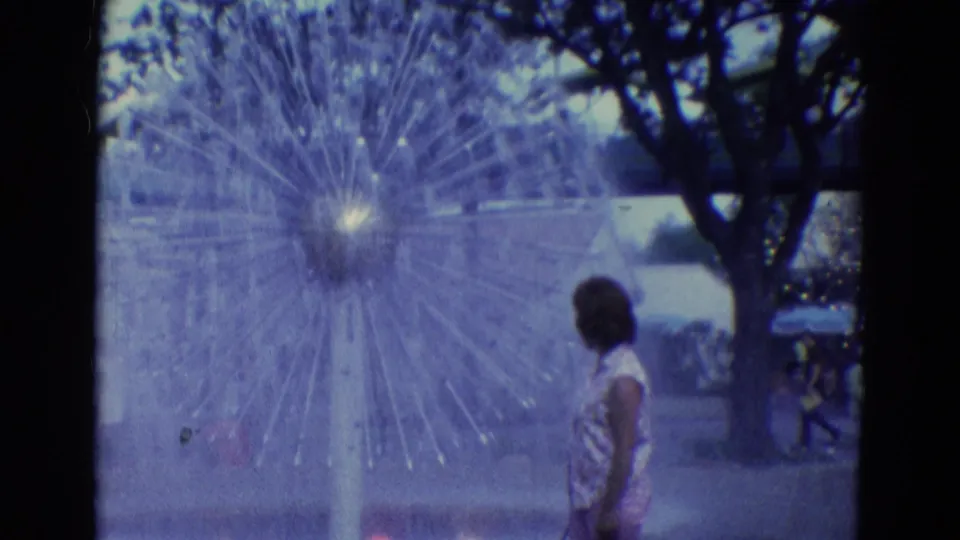 1969 Diller, Nebraska 8mm frame emphasizing the fountain spray pattern with a woman standing nearby under trees.