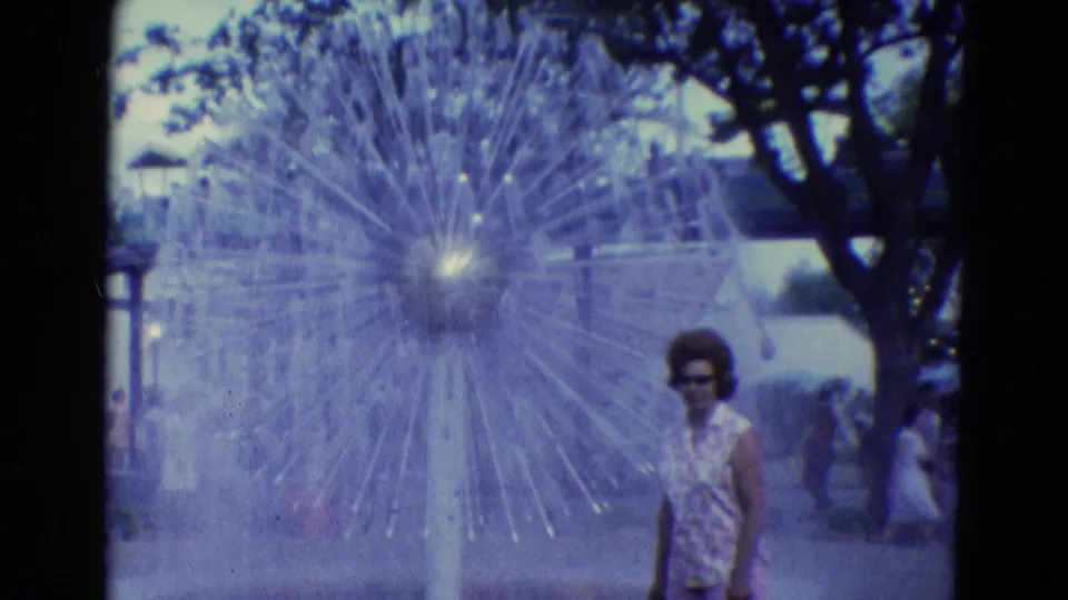 1969 Diller, Nebraska frame showing a woman in sunglasses passing the large fountain in a public tree-lined setting.