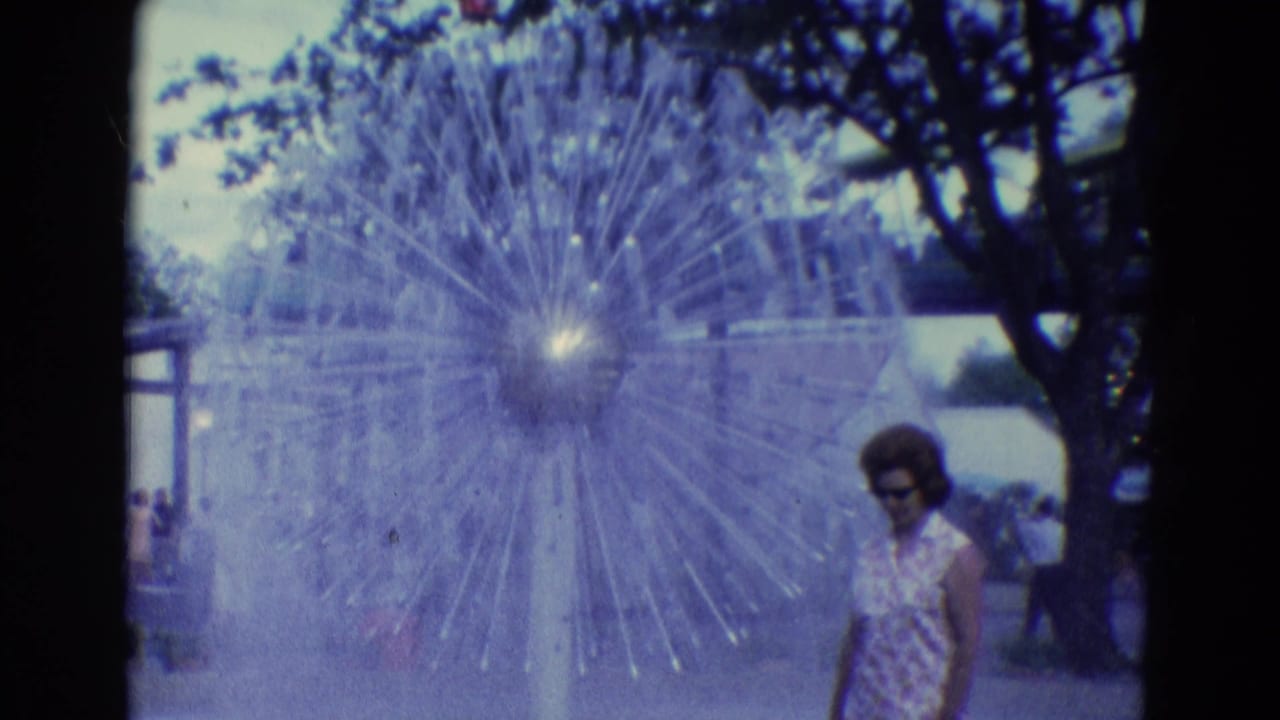 In Diller, Nebraska, 1969, a woman in sunglasses and pink pants stands near a large radial fountain in an outdoor public area.
