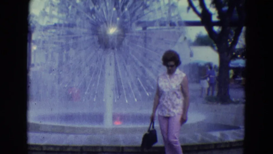 A woman in pink pants and sunglasses walks near the large fountain in the supplied 1969 Diller, Nebraska home-movie scene.