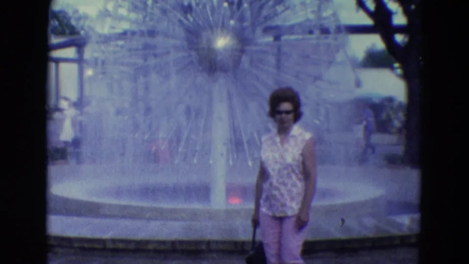 The woman faces the camera near the starburst fountain, with the fountain basin, trees, and distant visitors visible behind her.