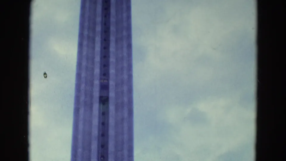 The camera continues on the elevated lift tower, showing its vertical sides and central track against pale clouds.