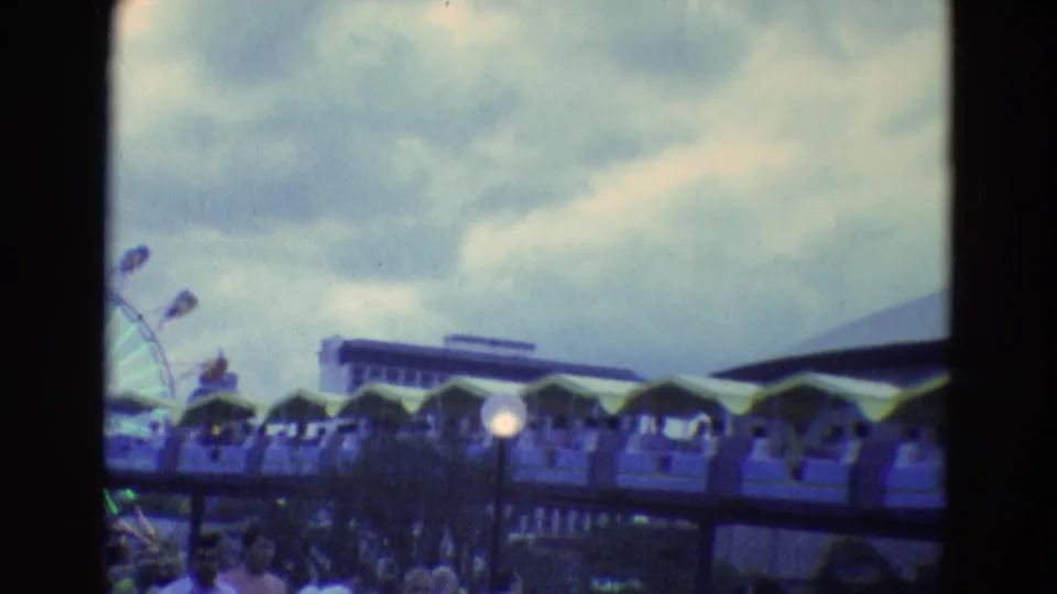 1969 8mm fairground frame showing covered ride cars, lamps, cloudy sky, and a partial Ferris wheel in the distance.