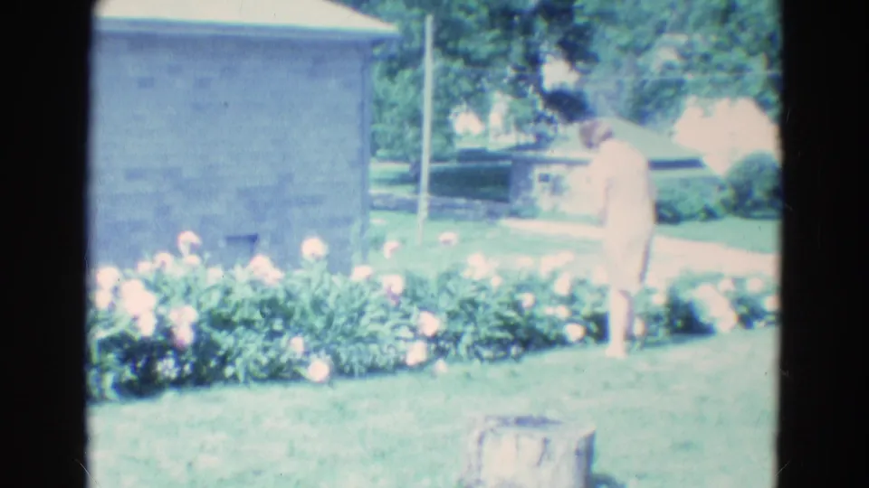 1969 Diller, Nebraska home-movie frame showing a woman in a pale dress walking across a sunlit lawn toward the flower bed.