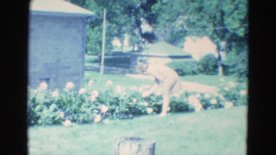 1969 Diller, Nebraska 8mm frame showing a woman bending toward a blooming flower bed in a bright residential-looking yard.
