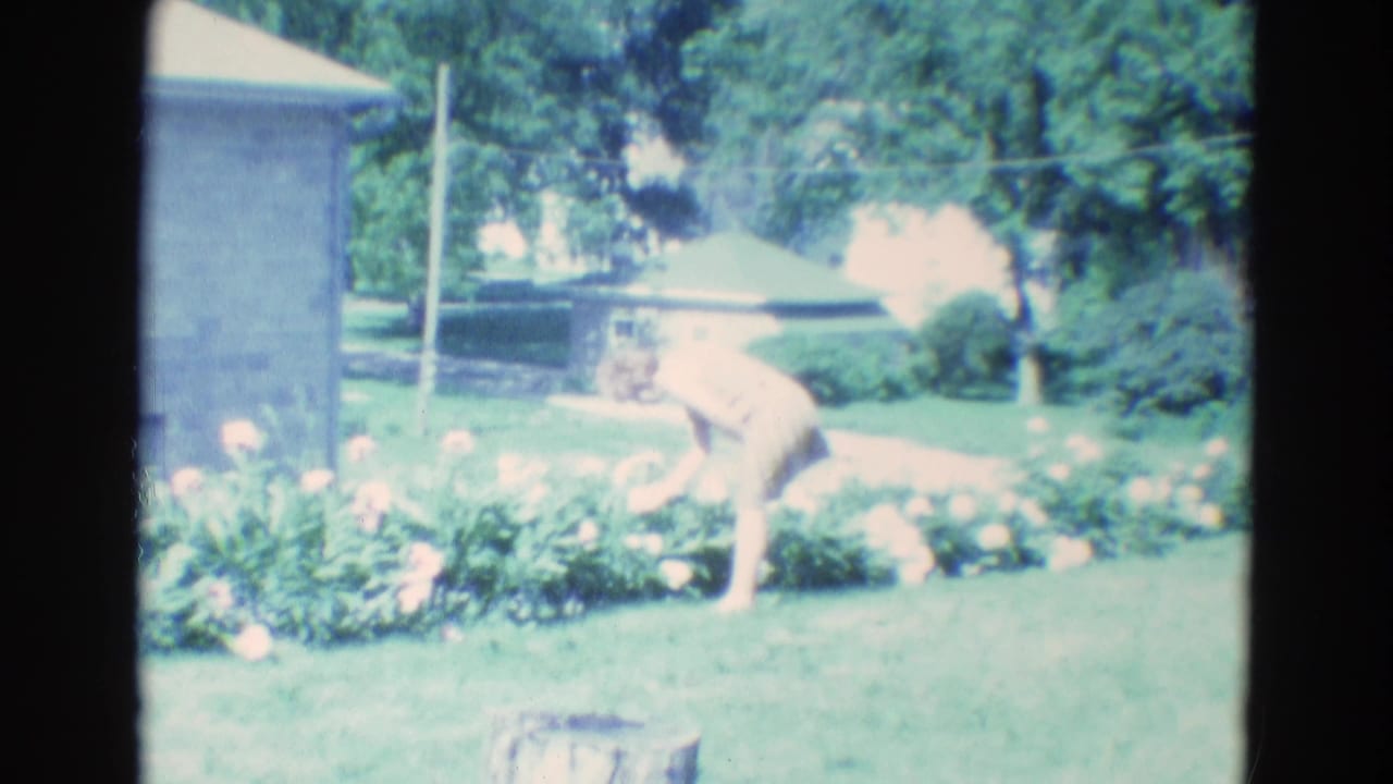In Diller, Nebraska 1969 home-movie footage, a woman in a light dress bends toward flowers beside a lawn and low house.
