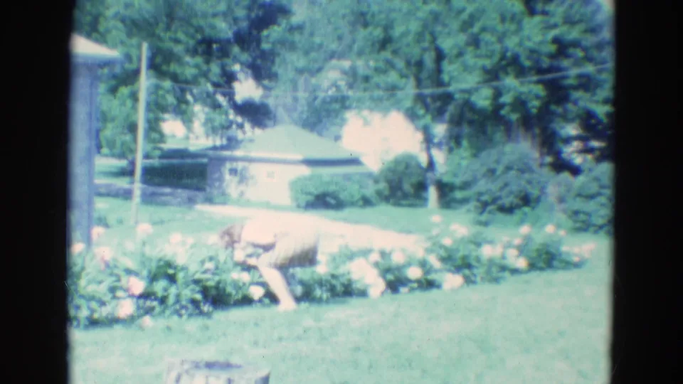 The woman crouches close to a flower bed in the 1969 Diller, Nebraska yard, with trees and a low house behind her.