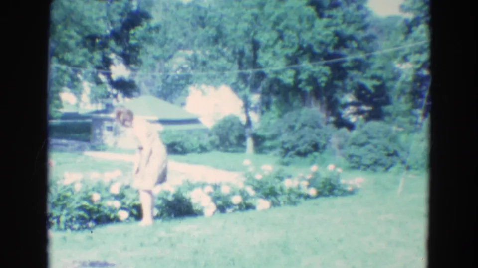 The woman stands beside the flowers in a Diller, Nebraska residential yard, with the soft color and focus of 1969 8mm footage.