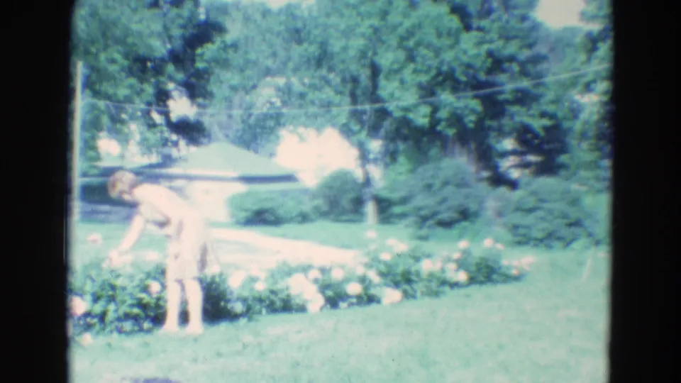 At the end of the clip, the woman bends toward the flowers in the 1969 Nebraska yard, framed by lawn, trees, and a low house.