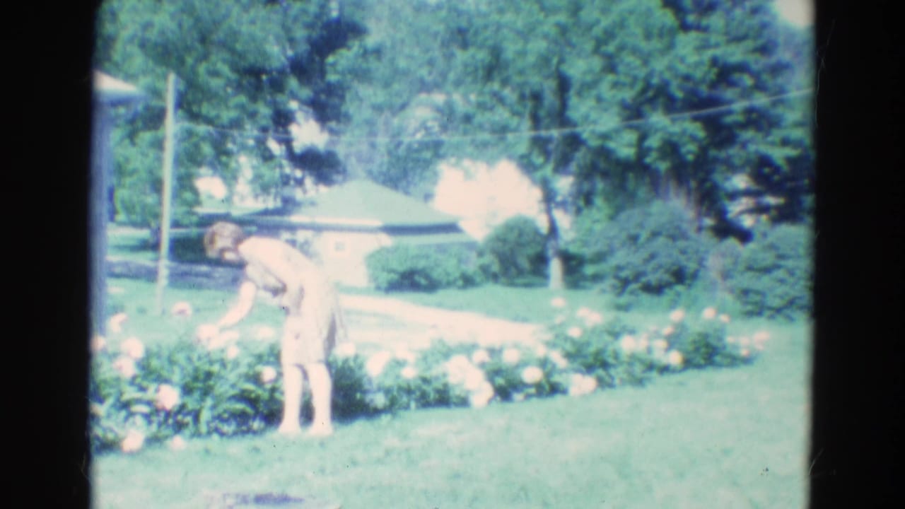 Woman bends over a flower bed in a green yard near trees and a house.