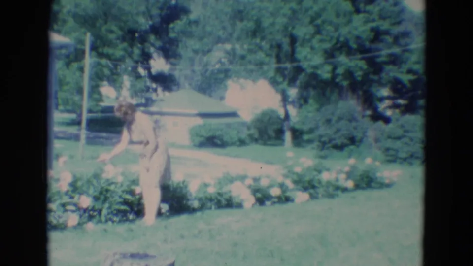 The woman stands upright near the garden bed in a small-town Nebraska yard, with trees and shrubs behind her.