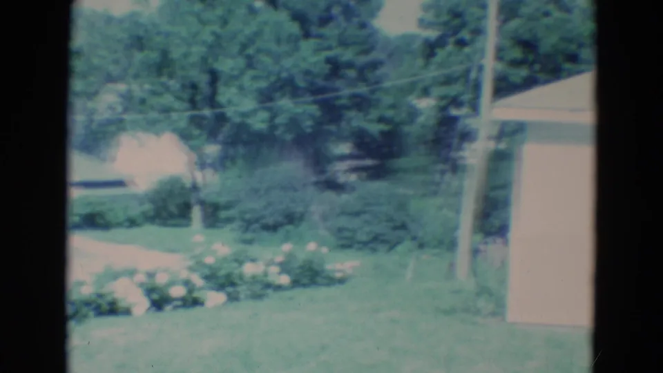 The woman has moved out of frame, leaving the flower bed, lawn, trees, and house corner visible in the 1969 yard scene.