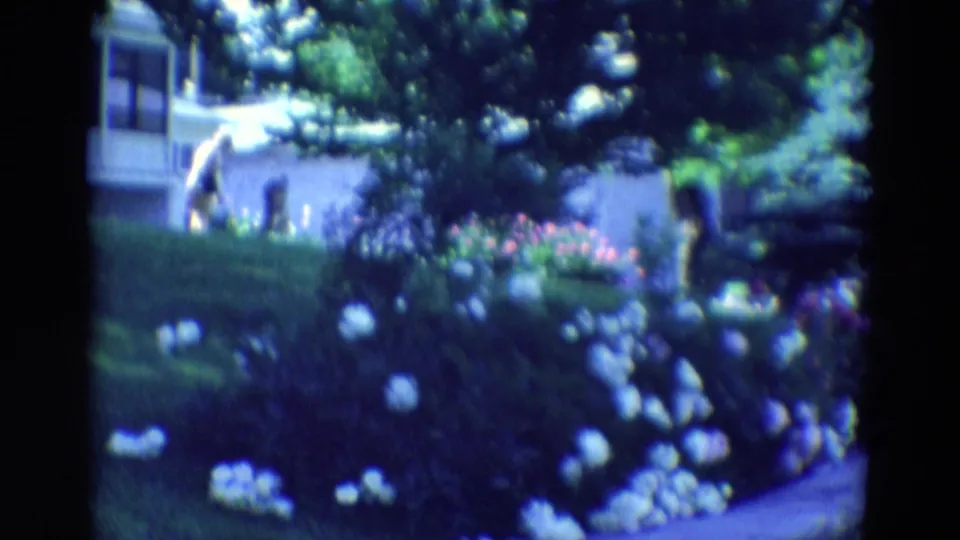 1969 Diller, Nebraska frame with a person briefly bending near a residential garden beside a light house and flower beds.
