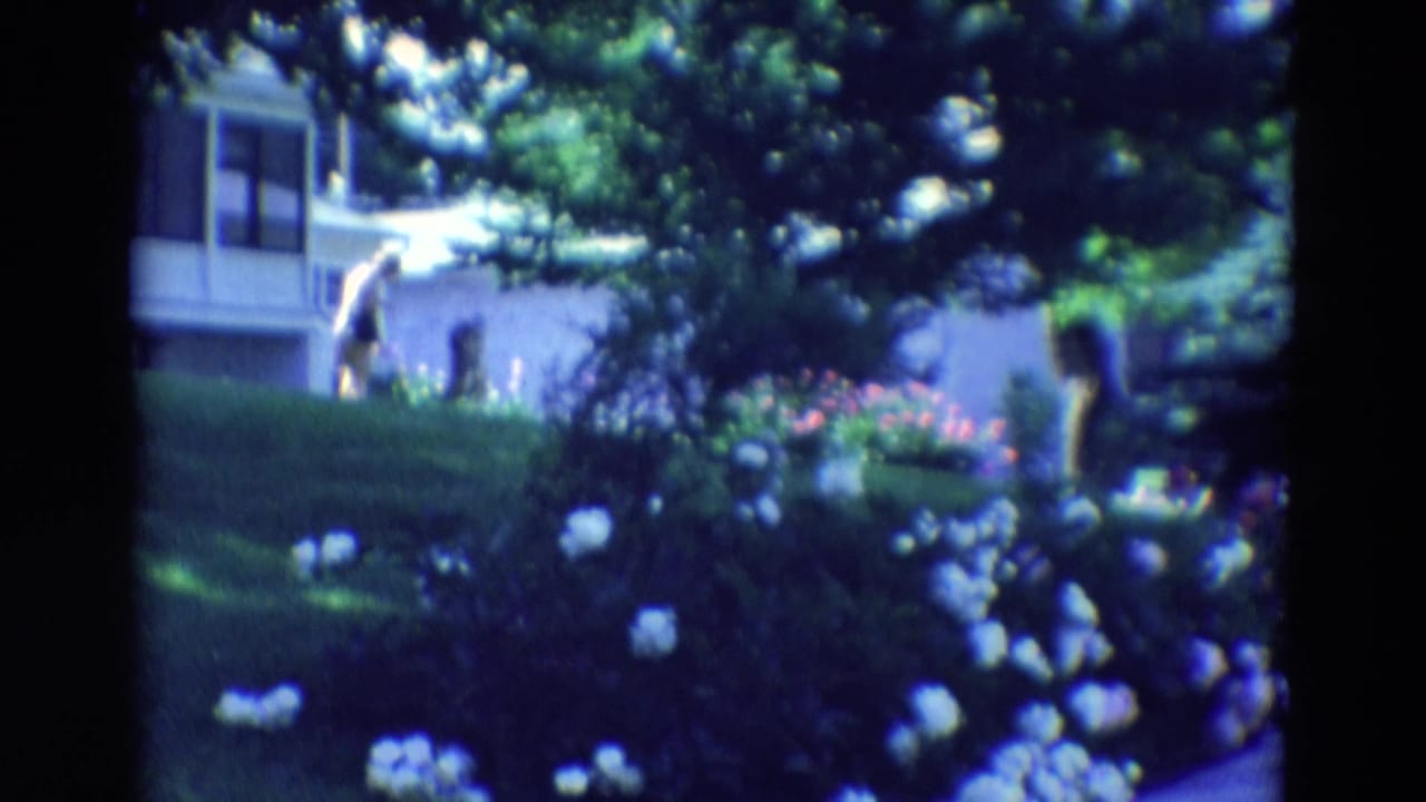 Person in light clothing walking across a lawn near a white house and flowerbeds.